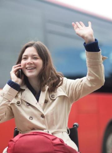 Jeune femme en fauteuil roulant faisant un signe de la main depuis une station de bus.